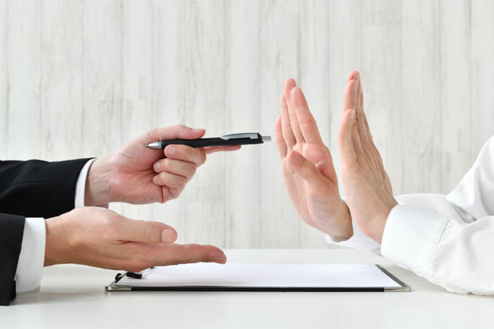 Twee paar handen van personen tegenover elkaar. Tussen de personen in ligt een document. De handen van de persoon aan de linker zijde overhandigen een pen en de persoon aan de rechter zijde wijst af door zijn handen op te steken.