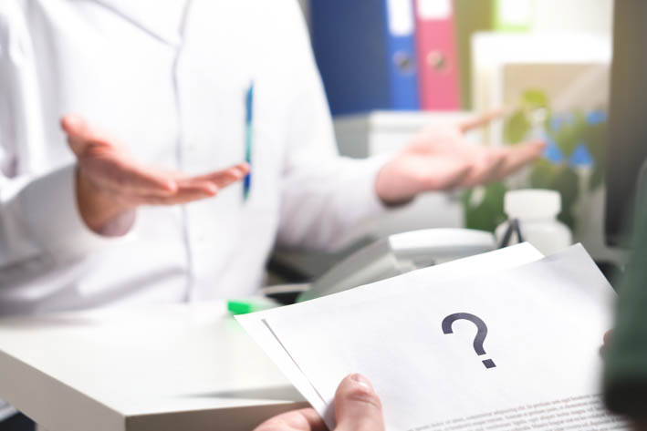 Persoon in een wit overhemd die aan een bureau zit, beide handen opsteekt en een vragende houding uitstraalt. Een andere persoon houdt een stuk papier vast met een groot vraagteken erop.