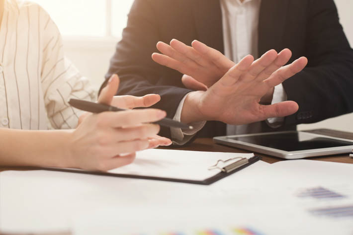 Twee mannen die aan een tafel zitten en in gesprek zijn. Eén man houdt een pen vast en lijkt aantekeningen te maken op een klembord, terwijl de andere man zijn handen heeft opgeheven in een gebaar dat kan duiden op stoppen.
