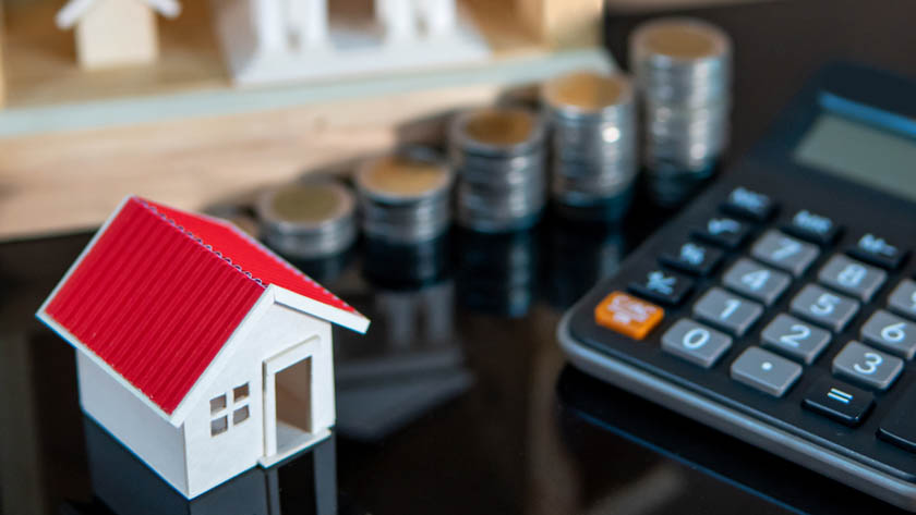 Miniatuurhuis en een rekenmachine, met op de achtergrond enkele stapels euromunten.