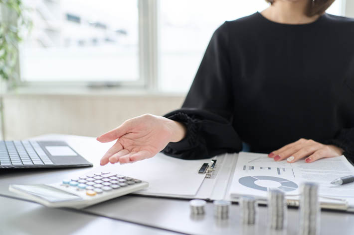 Vrouw die aan een bureau zit met verschillende kantoorartikelen. De vrouw maakt een gebaar met één hand terwijl de andere hand op enkele documenten rust. Op het bureau staan een open laptop, een rekenmachine, stapels munten en documenten.