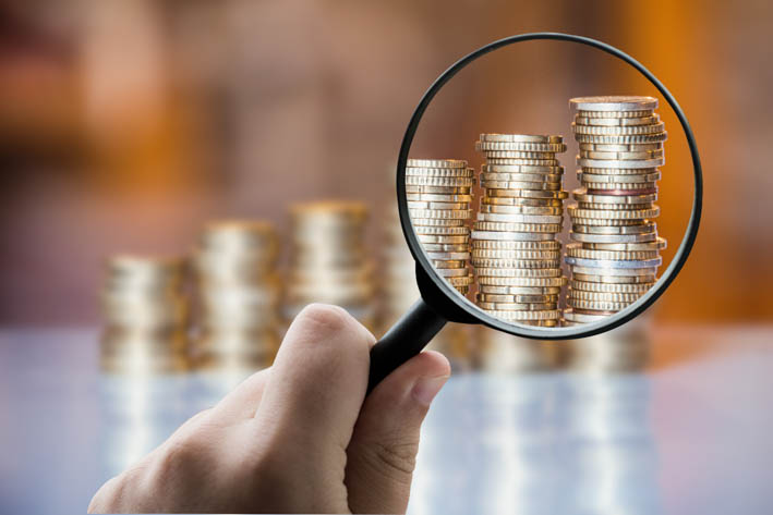 Hand met vergrootglas. In het kader van het glas is een stapel munten helder zichtbaar. Op de achtergrond zijn stapels met munten wazig zichtbaar.