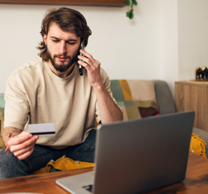 Een persoon zit op een bank en houdt een bankpas in de ene hand terwijl die met de andere hand telefoneert. Op de voorgrond staat een open laptop. De persoon kijkt verbaast naar de bankpas.