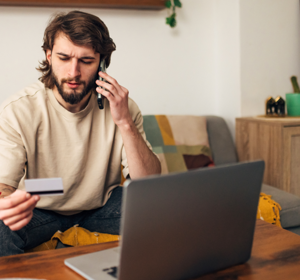 Een persoon zit op een bank en houdt een bankpas in de ene hand terwijl die met de andere hand telefoneert. Op de voorgrond staat een open laptop. De persoon kijkt verbaast naar de bankpas.