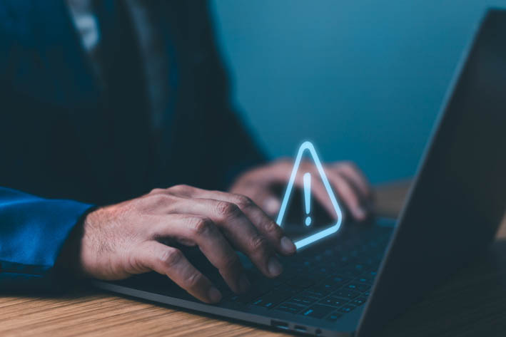 Persoon die op een laptoptoetsenbord typt, met een verlicht waarschuwingsdriehoeksymbool (met een uitroepteken erin) dat boven het toetsenbord zweeft.