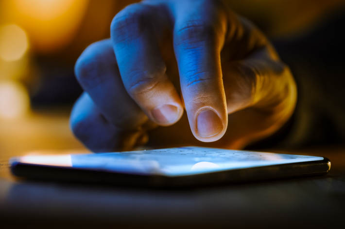 Close-up van een hand die een smartphone bedient, met blauw licht van het scherm in een donkere ruimte.