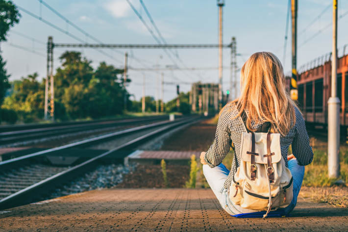 Achterkant van een vrouw met een rugzak om, die bij een treinstation zit. Ze kijkt richting een leeg treinspoor.