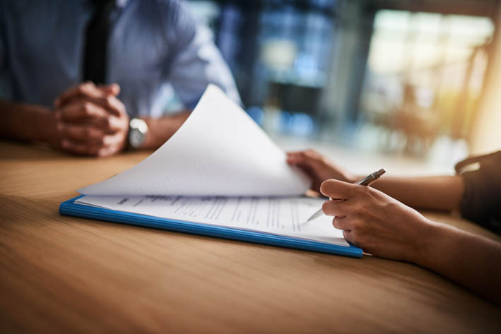 Twee personen die aan een tafel zitten, waarbij één persoon een pen vasthoudt en bezig lijkt te zijn met het ondertekenen of beoordelen van een document.