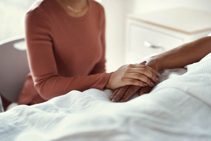 Een persoon die naast een bed zit en de hand vasthoudt van een andere persoon die in het bed ligt.