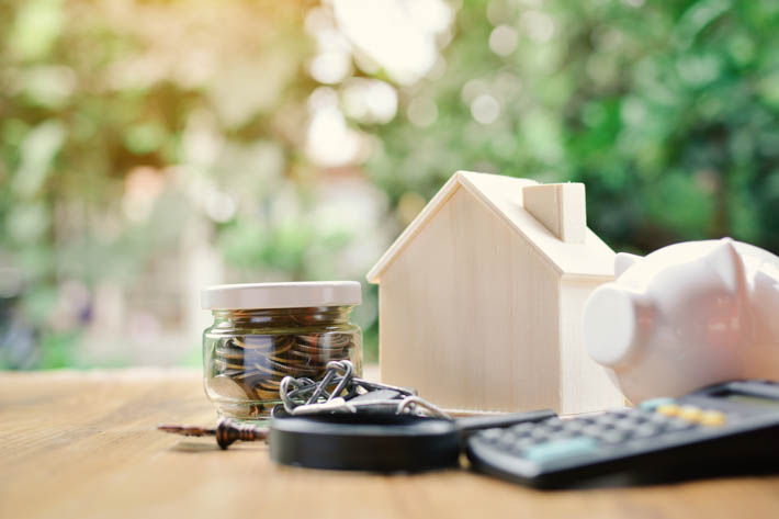 Bureau met pot geld, houten huis, spaarvarken, rekenmachine en kettingslot. De achtergrond is een natuurlijke omgeving met veel groen. 
