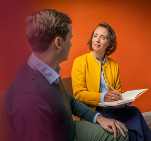 Een man en een vrouw die tegenover elkaar zitten op een bank. De vrouw glimlacht en heeft een notitieboekje en een een pen in haar hand.