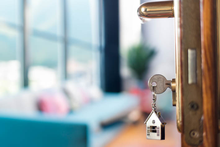 Close-up van een sleutel die in een deurslot is gestoken. Aan de sleutel hangt een kleine sleutelhanger in de vorm van een huis. Op de achtergrond is een onscherpe weergave een interieur.