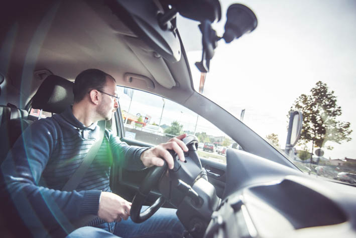 Een man zit in een auto en kijkt naar links terwijl hij het stuur vasthoudt. De zon schijnt fel door het raam aan de bestuurderskant.