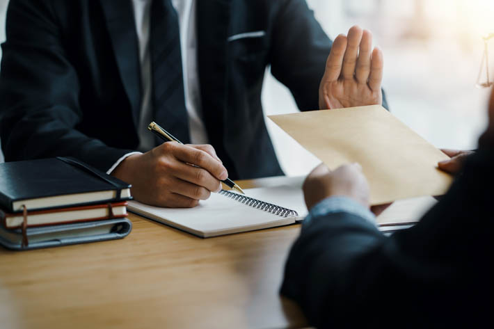 Twee personen in een formele setting. Eén persoon overhandigt een envelop aan de ander, die een hand opsteekt alsof ze de actie willen weigeren of stoppen.