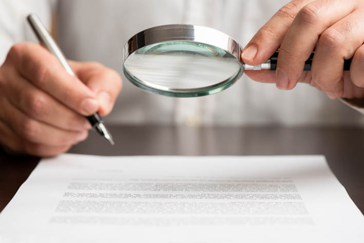 Een man houdt een vergrootglas en een pen vast terwijl hij een document met kleine tekst bekijkt.