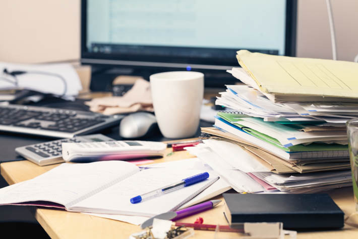Rommelig bureau met documenten, een rekenmachine, een kopje en een computer.