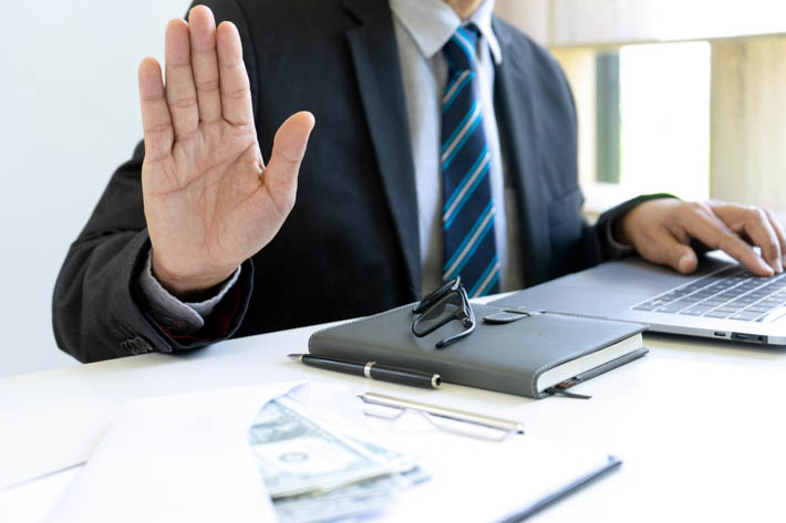 Een man in een pak gebaart met zijn hand 'stop'. Op het bureau voor hem is een laptop zichtbaar en enkele kantoorartikelen.