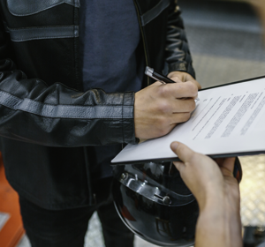 Een persoon in een zwart leren jack ondertekent een document op een clipboard, terwijl een andere persoon het vasthoudt. Op de voorgrond is een motorhelm zichtbaar die wordt vastgehouden. 