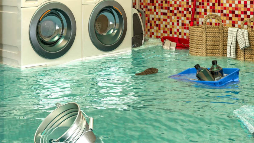 Wasruimte die onder water staat. Verschillende items zoals een metalen emmer, een blauwe plastic container met flessen en gevlochten manden drijven of zijn gedeeltelijk ondergedompeld in het water.