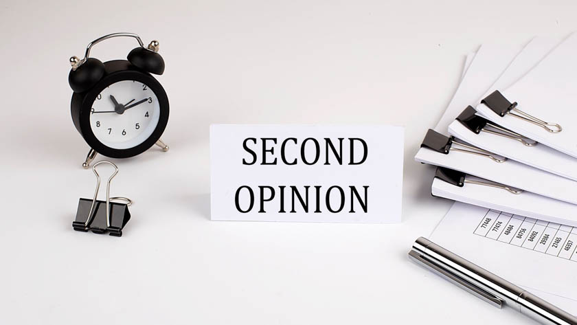 Zwarte wekker, verschillende stapels papieren die met zwarte binders zijn vastgeklikt, een pen en een kaart met de tekst "SECOND OPINION" erop. 