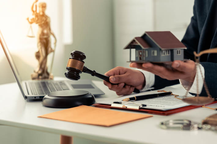 Een bureau met een laptop, documenten, een weegschaal en een persoon die een gavel en een modelhuis vasthoudt.