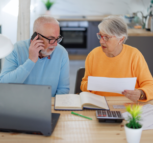 Een oudere man en vrouw zitten samen aan een keukentafel met een laptop en papieren. De man belt met zijn telefoon en kijkt bezorgd, terwijl de vrouw een document vasthoudt en hem vragend aankijkt.