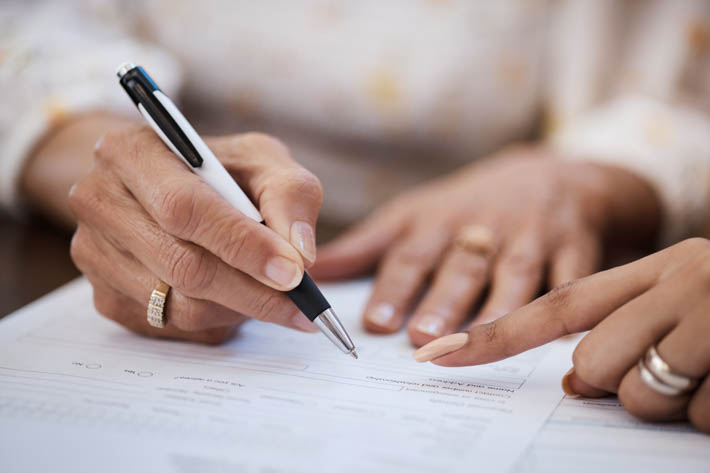 Twee handen, waarbij één hand een pen vasthoudt en op een stuk papier schrijft, terwijl de andere hand naar het papier wijst.