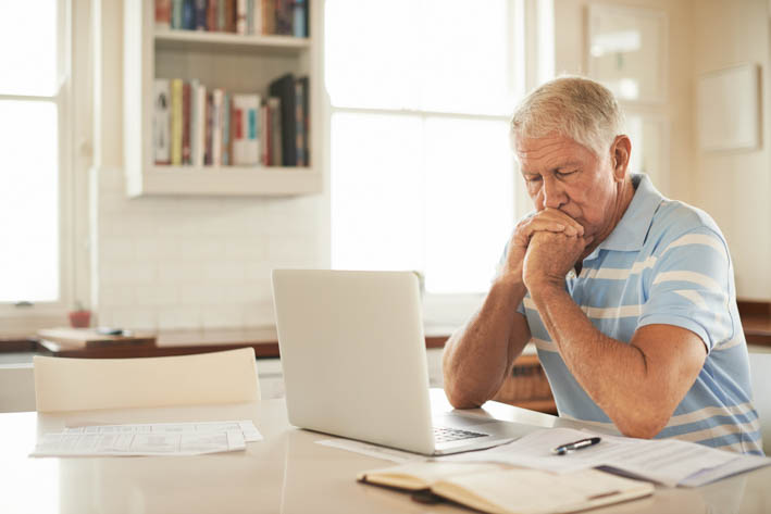 Oudere man die aan een tafel zit. Op de tafel staat een laptop, met daarnaast een open notitieblok en enkele documenten. De man heeft rust zijn hoofd op zijn handen en straalt uit na te denken.