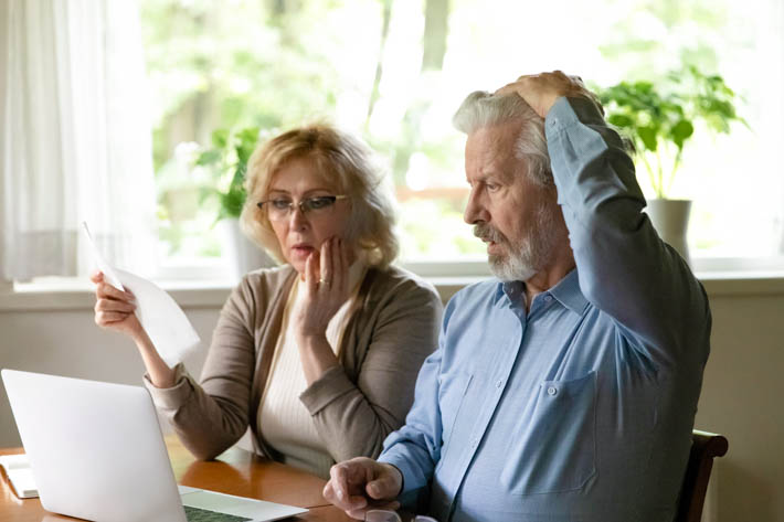 Ouder stel zit aan tafel met een laptop; de vrouw houdt een document vast terwijl ze bezorgd kijkt, en de man raakt met zijn hand zijn hoofd aan.