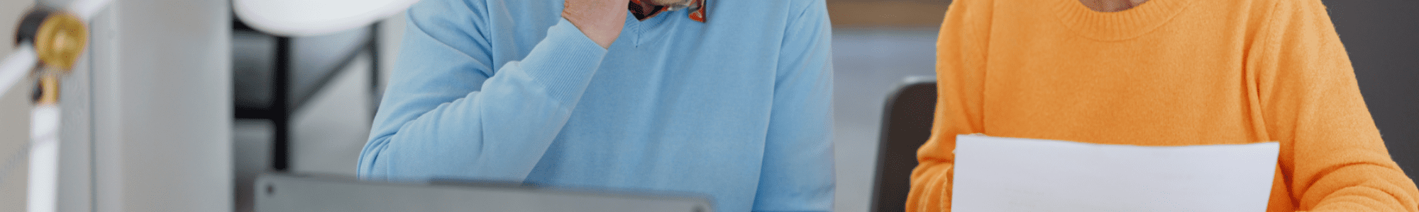 Een ouder echtpaar zit aan een tafel met een laptop, papieren, een notitieboek en een rekenmachine. De man belt terwijl de vrouw een document vasthoudt en informatie bespreekt.