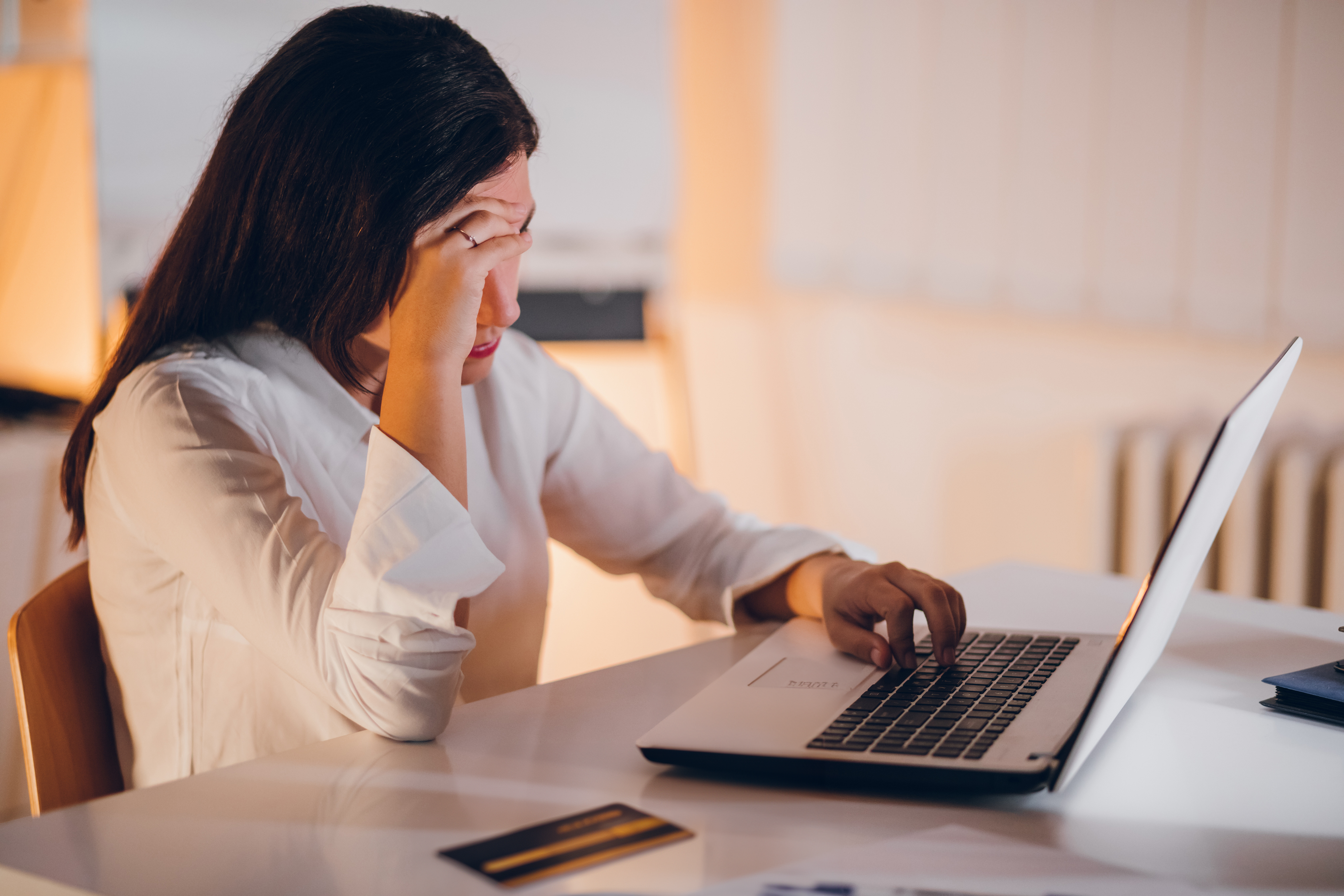 Vrouw zit achter bureau en kijkt zorgelijk naar laptop met hand voor haar gezicht. Op tafel licht een bankpas.
